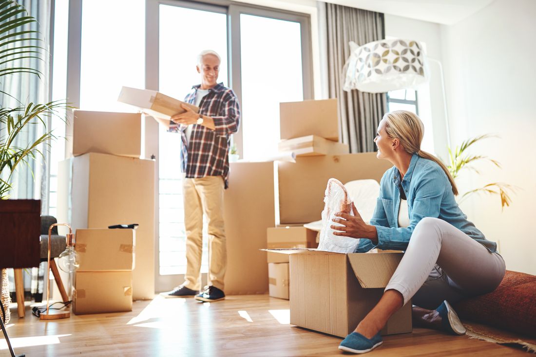 Gray-haired man and blond woman unpacking moving boxes in a bright room with tall windows.