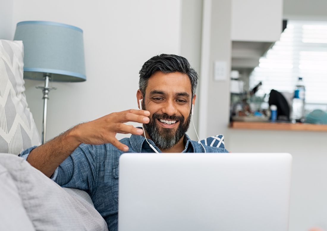 Smiling bearded man wearing earphones on a video call, laptop on his lap, sitting on a sofa at home.